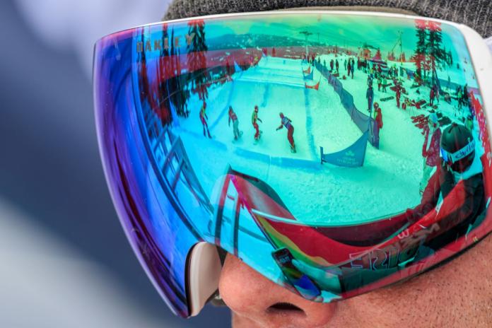 A close up shot of an athlete's goggles, which shows the reflection of snowboarding competition